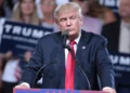 Donald Trump berdiri di belakang podium dan mikrofon dengan jas gelap serta dasi merah di depan peserta kampanye