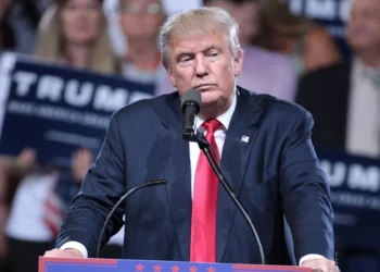 Donald Trump berdiri di belakang podium dan mikrofon dengan jas gelap serta dasi merah di depan peserta kampanye