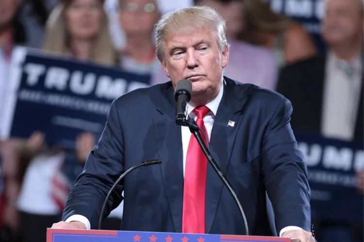 Donald Trump berdiri di belakang podium dan mikrofon dengan jas gelap serta dasi merah di depan peserta kampanye