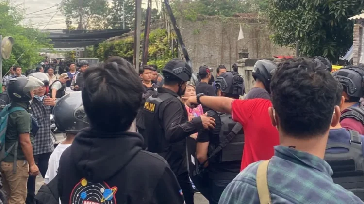 Sejumlah aparat kepolisian berhelm dan mengenakan perlengkapan pengamanan berada di antara warga di sebuah jalan permukiman