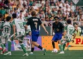 Pemain Barcelona berseragam gelap menggiring bola menghadapi pemain Real Betis berseragam hijau-putih di lapangan.