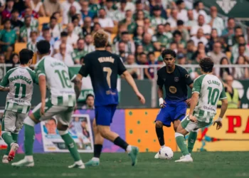 Pemain Barcelona berseragam gelap menggiring bola menghadapi pemain Real Betis berseragam hijau-putih di lapangan.