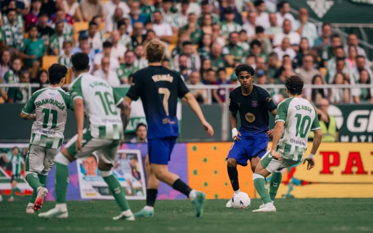Pemain Barcelona berseragam gelap menggiring bola menghadapi pemain Real Betis berseragam hijau-putih di lapangan.