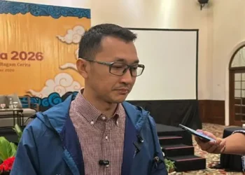 Seorang pria berkacamata mengenakan jaket biru sedang diwawancarai oleh wartawan di dalam ruangan
