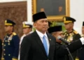 Seorang pria mengenakan jas hitam, dasi biru dan peci hitam berdiri di dekat mikrofon, dengan sejumlah perwira berseragam di belakangnya