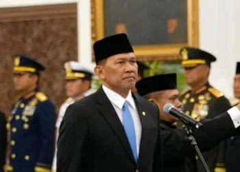 Seorang pria mengenakan jas hitam, dasi biru dan peci hitam berdiri di dekat mikrofon, dengan sejumlah perwira berseragam di belakangnya