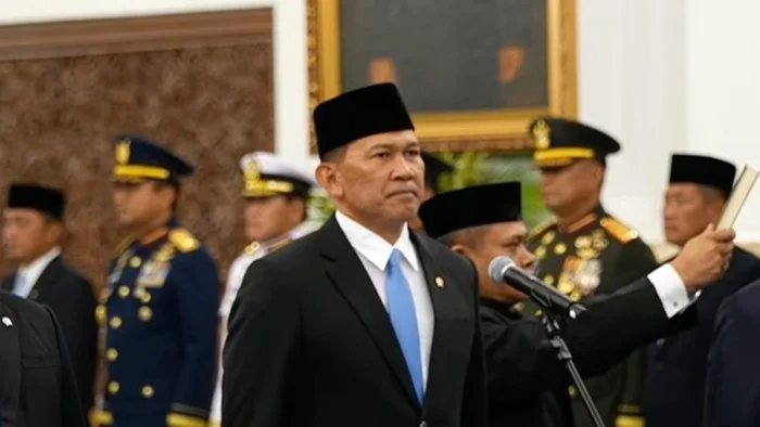 Seorang pria mengenakan jas hitam, dasi biru dan peci hitam berdiri di dekat mikrofon, dengan sejumlah perwira berseragam di belakangnya