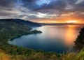 Pemandangan danau yang luas dengan perbukitan hijau di tepi danau serta langit jingga saat matahari terbenam