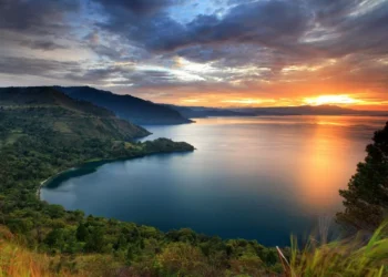 Pemandangan danau yang luas dengan perbukitan hijau di tepi danau serta langit jingga saat matahari terbenam