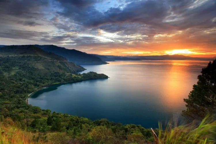 Pemandangan danau yang luas dengan perbukitan hijau di tepi danau serta langit jingga saat matahari terbenam