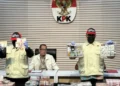 Dua petugas KPK memperlihatkan uang dan barang bukti lain dalam konferensi pers di depan lambang KPK, dengan sejumlah bundel uang di atas meja