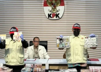 Dua petugas KPK memperlihatkan uang dan barang bukti lain dalam konferensi pers di depan lambang KPK, dengan sejumlah bundel uang di atas meja