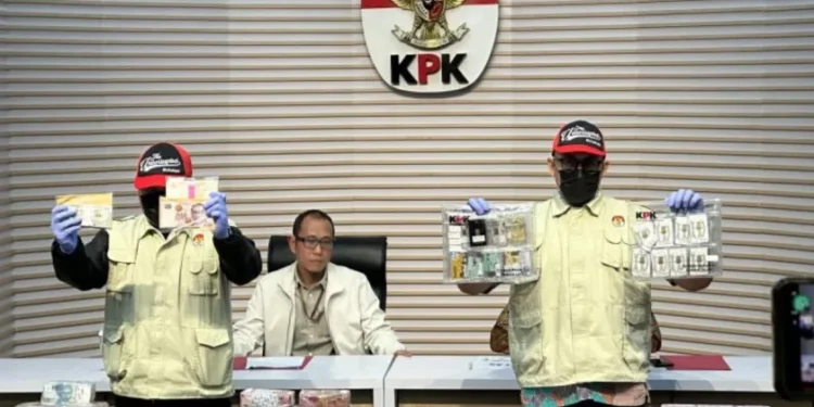 Dua petugas KPK memperlihatkan uang dan barang bukti lain dalam konferensi pers di depan lambang KPK, dengan sejumlah bundel uang di atas meja