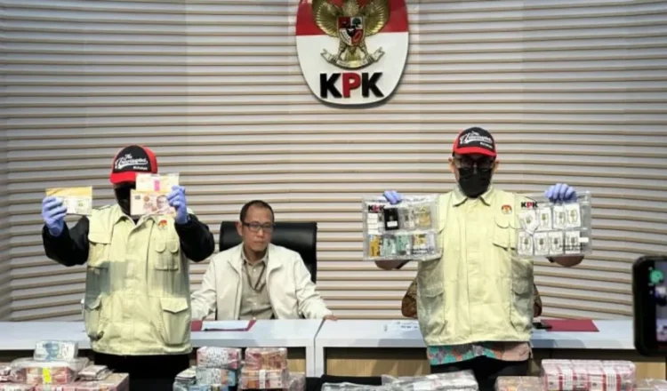 Dua petugas KPK memperlihatkan uang dan barang bukti lain dalam konferensi pers di depan lambang KPK, dengan sejumlah bundel uang di atas meja