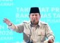 Presiden Prabowo Subianto mengenakan kemeja krem dan peci hitam berbicara di depan mikrofon sambil mengangkat tangan