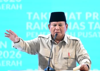 Presiden Prabowo Subianto mengenakan kemeja krem dan peci hitam berbicara di depan mikrofon sambil mengangkat tangan