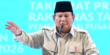 Presiden Prabowo Subianto mengenakan kemeja krem dan peci hitam berbicara di depan mikrofon sambil mengangkat tangan
