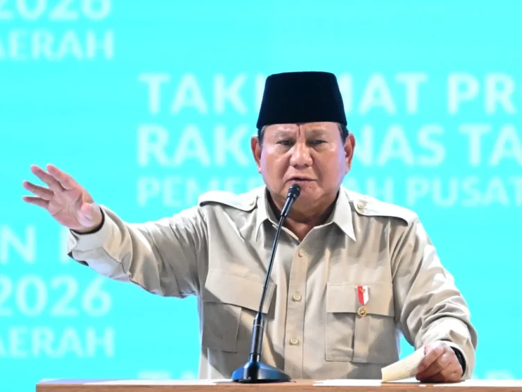 Presiden Prabowo Subianto mengenakan kemeja krem dan peci hitam berbicara di depan mikrofon sambil mengangkat tangan