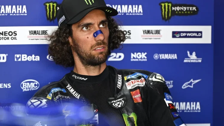 Alex Rins mengenakan baju balap Monster Energy Yamaha dan topi hitam di depan latar sponsor Yamaha