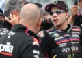 Marco Bezzecchi mengenakan seragam balap Aprilia Racing dan berbicara dengan sejumlah anggota tim.