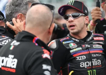 Marco Bezzecchi mengenakan seragam balap Aprilia Racing dan berbicara dengan sejumlah anggota tim.