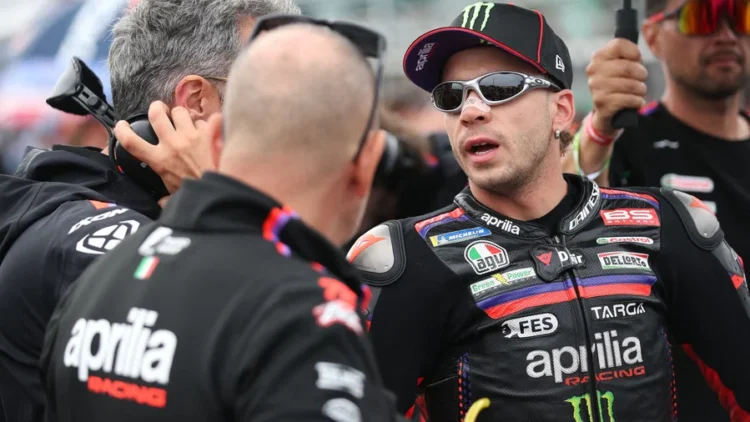 Marco Bezzecchi mengenakan seragam balap Aprilia Racing dan berbicara dengan sejumlah anggota tim.