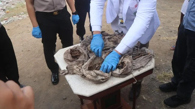 Petugas bersarung tangan biru memeriksa bungkusan kain kusam di atas meja, dikelilingi beberapa orang