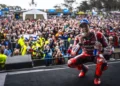 Marc Marquez memakai seragam balap Ducati merah dan topi biru, berjongkok sambil menggigit medali di depan penonton.