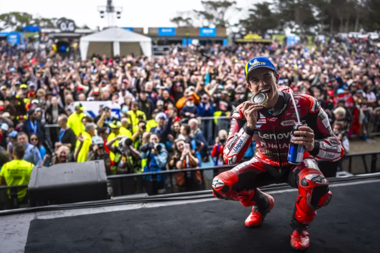 Marc Marquez memakai seragam balap Ducati merah dan topi biru, berjongkok sambil menggigit medali di depan penonton.