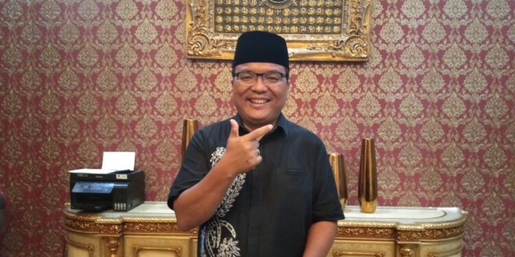 Denny Indrayana berdiri di dalam ruangan mengenakan peci hitam