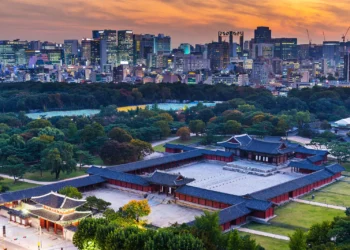 Pemandangan bangunan istana tradisional Korea dengan taman hijau dan gedung-gedung modern di belakangnya saat matahari terbenam