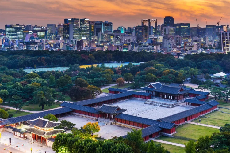 Pemandangan bangunan istana tradisional Korea dengan taman hijau dan gedung-gedung modern di belakangnya saat matahari terbenam