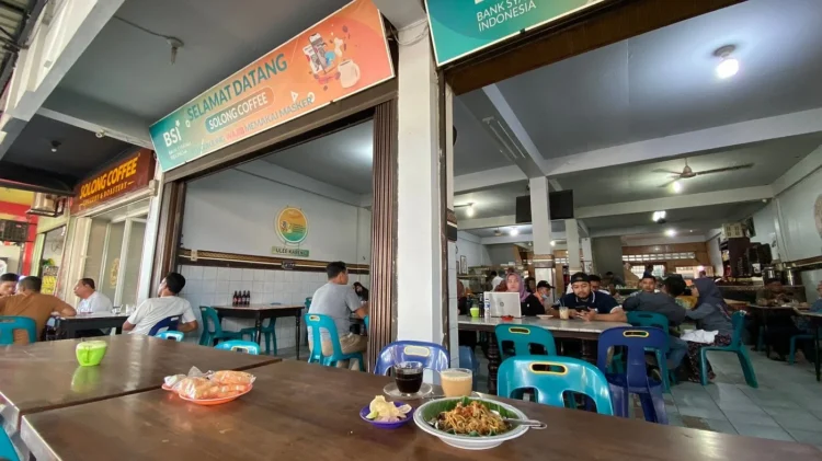 Suasana warung kopi Solong Coffee Ulee Kareng dengan pengunjung yang duduk di meja, serta minuman kopi dan hidangan di bagian depan.