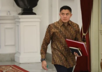 Seskab Teddy Indra Wijaya mengenakan kemeja batik dan membawa map merah saat berjalan di dalam gedung