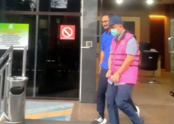 Pria mengenakan rompi tahanan merah muda dan masker berjalan didampingi petugas