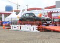 Mobil SUV berwarna hitam dengan logo Suzuki dipajang pada lintasan test drive berpalang merah, dengan tenda pameran di belakangnya.