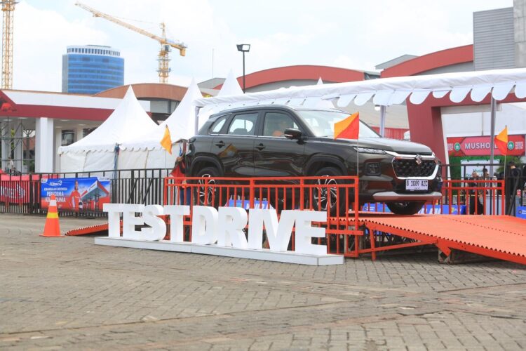 Mobil SUV berwarna hitam dengan logo Suzuki dipajang pada lintasan test drive berpalang merah, dengan tenda pameran di belakangnya.